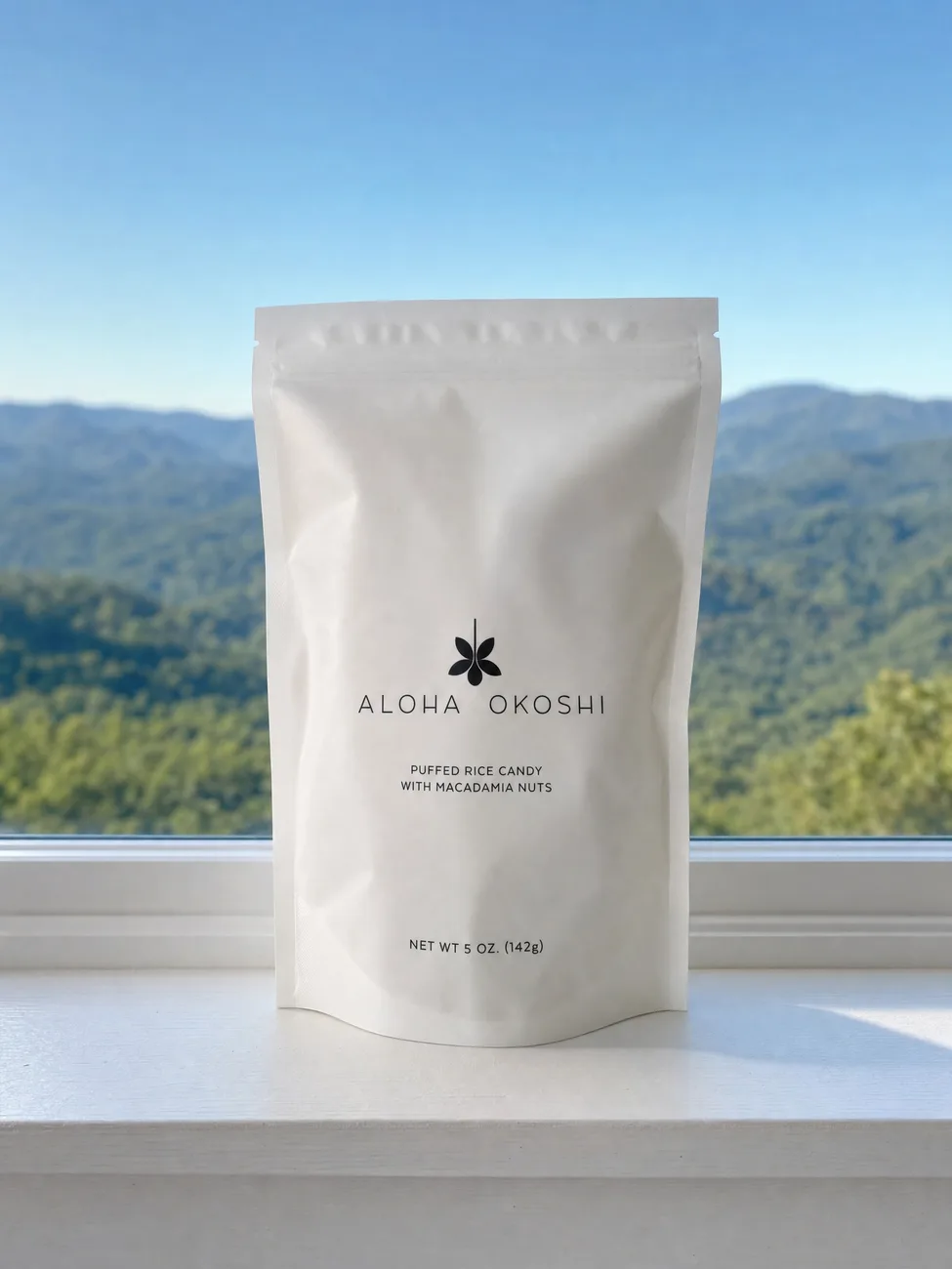 A bag of Aloha Okoshi puffed rice candy on a white windowsill with green mountains behind