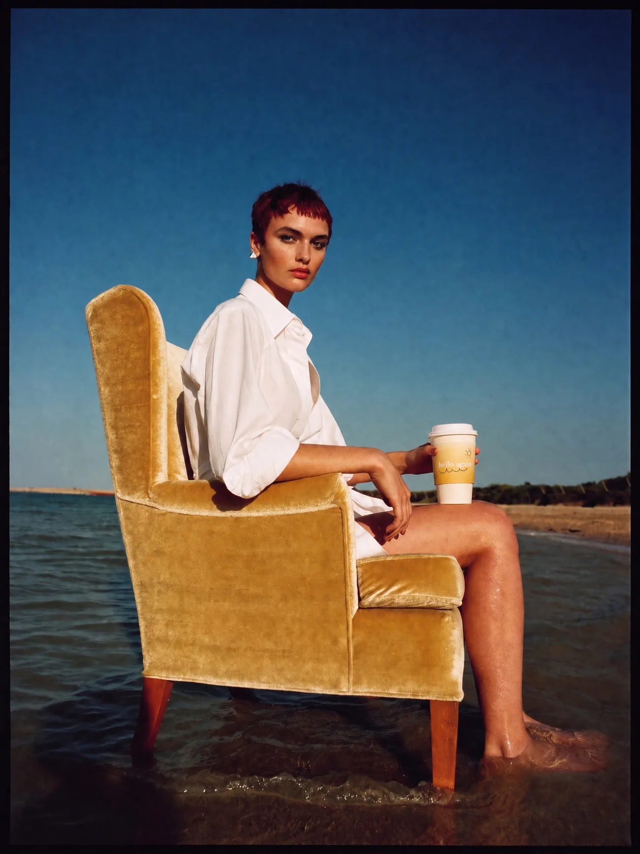 A woman in a gold velvet armchair in the surf holding a Hello Again cup