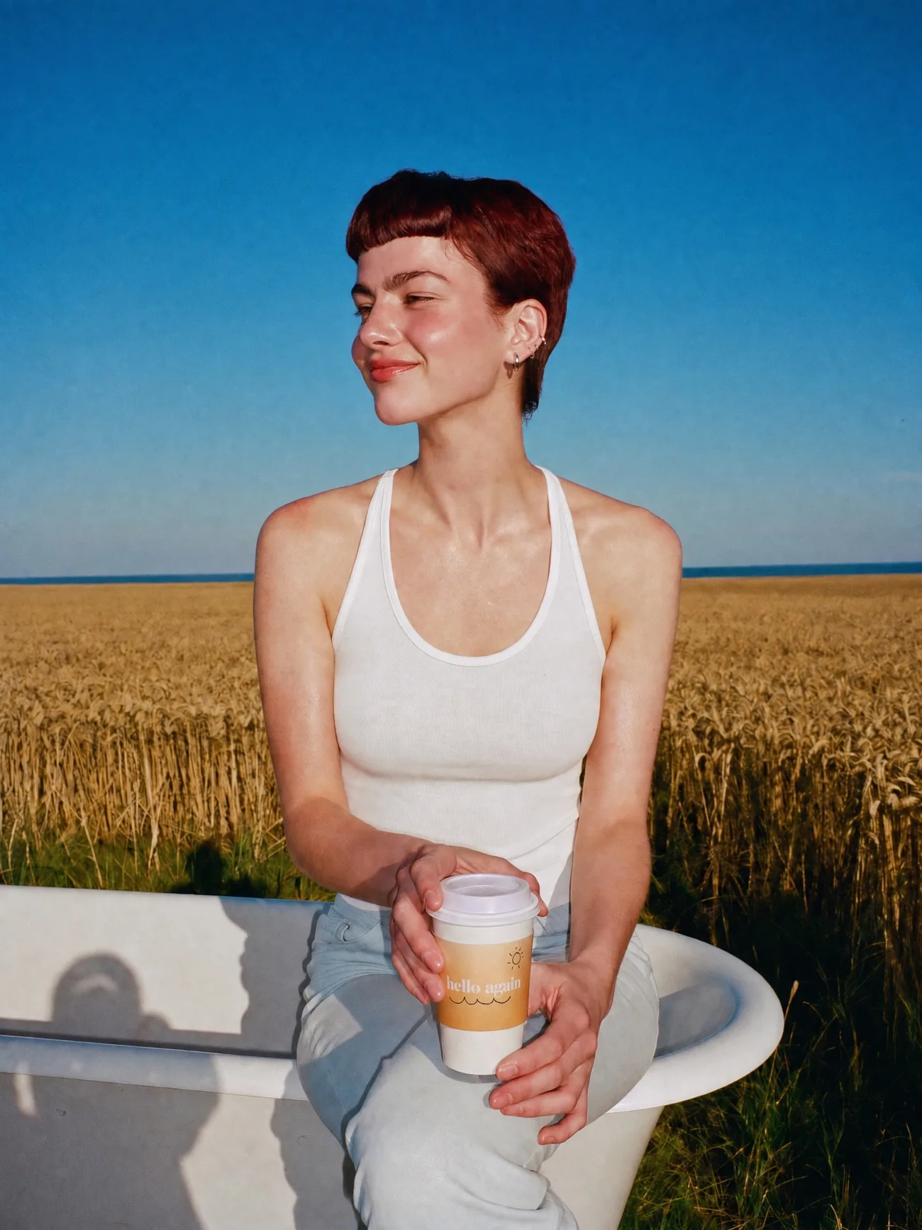 The same woman on a clawfoot bathtub in a wheat field with a Hello Again cup