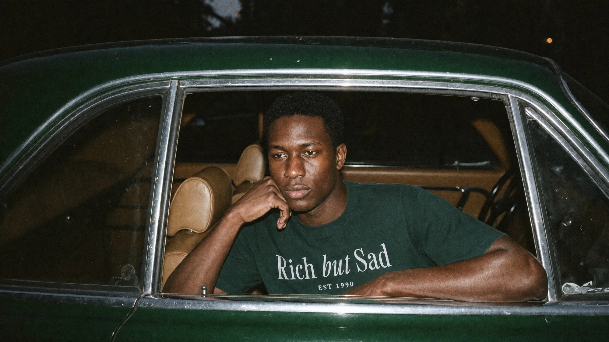 A man in the forest green Rich But Sad tee in the back seat of a racing green car at night