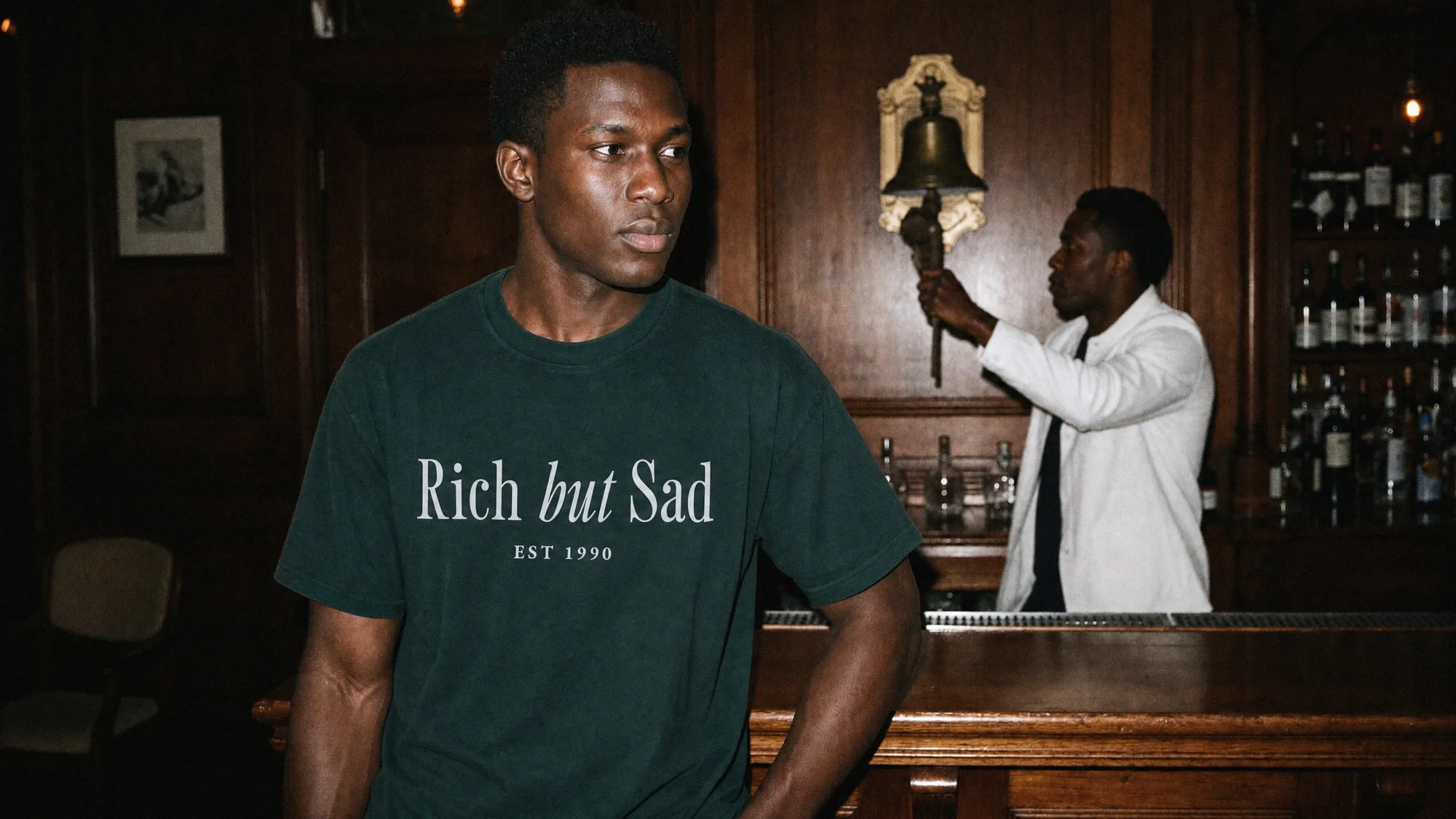 A man in the Rich But Sad tee at a wood-panelled bar while the barman rings the last orders bell