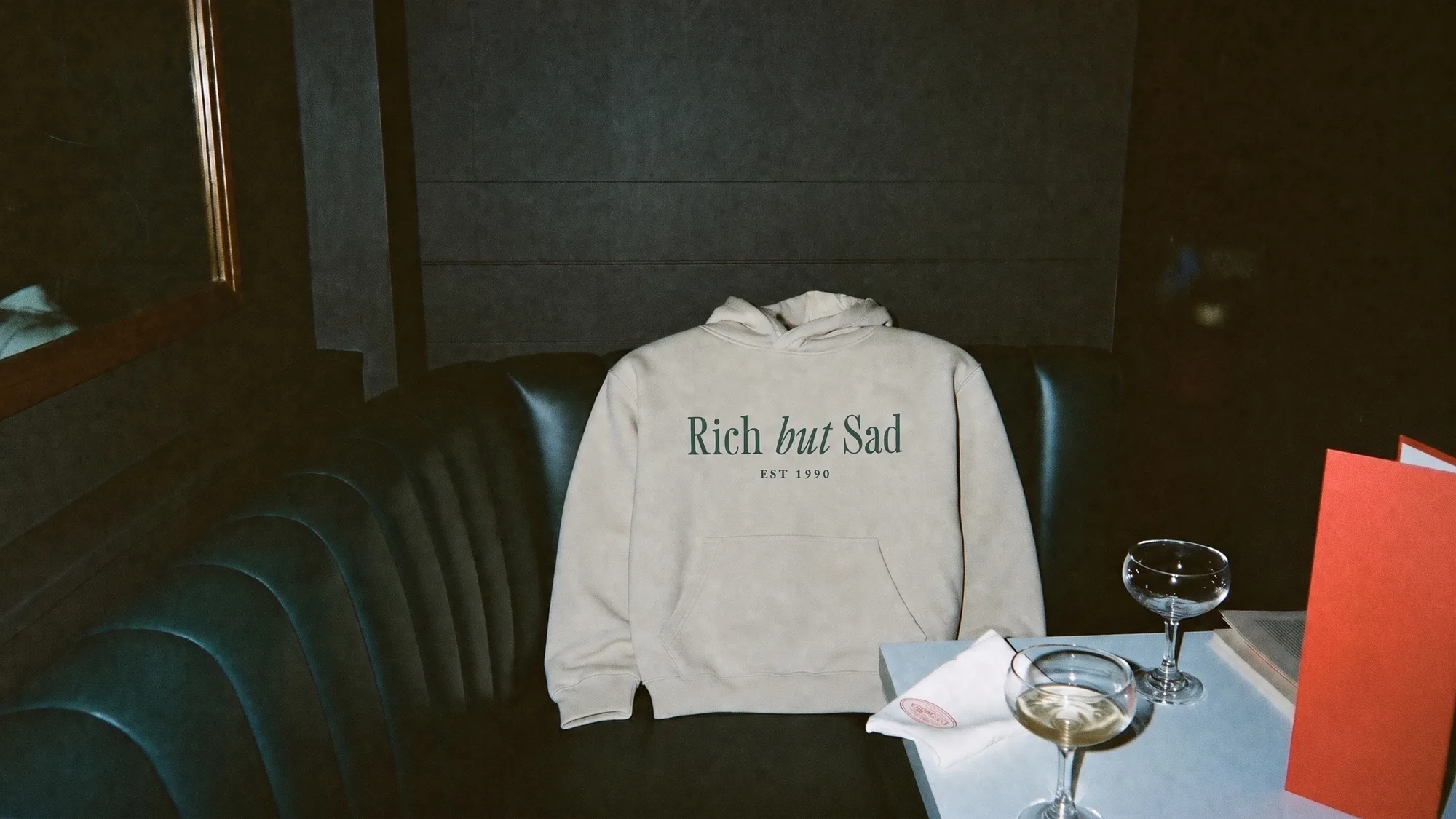 The oatmeal Rich But Sad hoodie left on a green leather banquette after closing